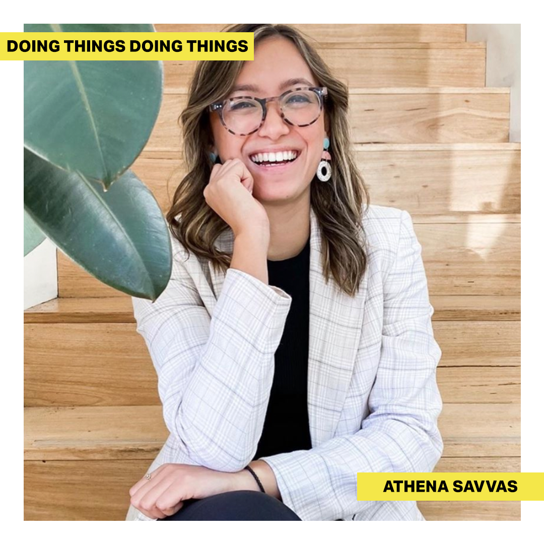 Athena Savvas sitting on wooden stairs and smiling widely, with the green leaves of a plant in the top right corner of image.